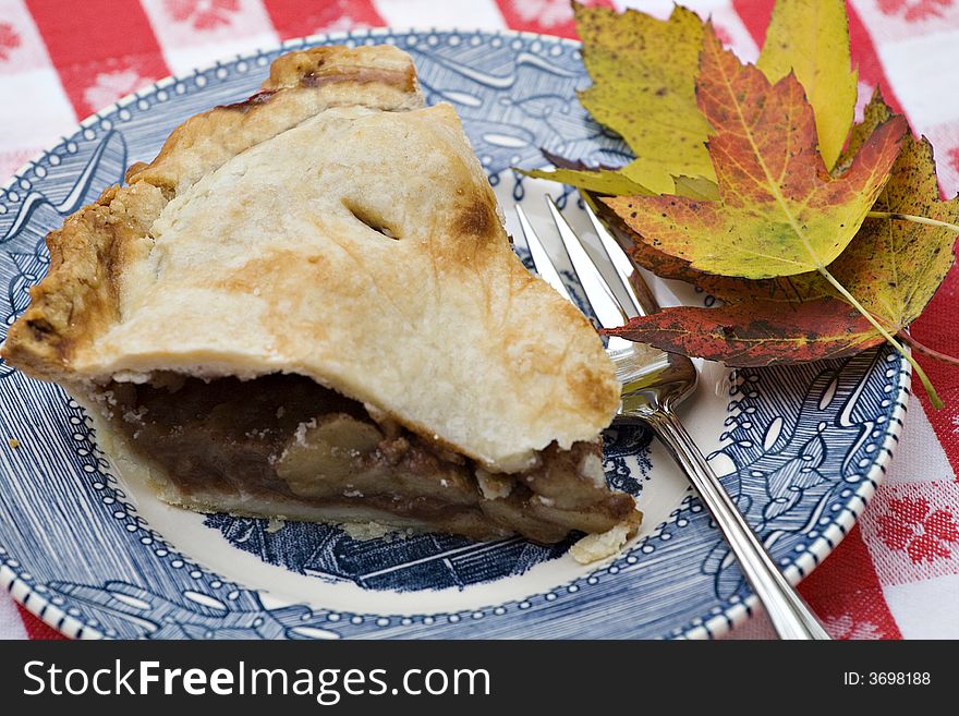 Slice of apple pie on old blue china and  gingham tablecloth.  Fall leaves on side. Slice of apple pie on old blue china and  gingham tablecloth.  Fall leaves on side.