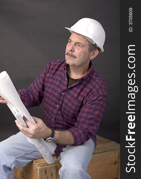 Middle-aged man with hardhat on holding blueprints in his hands while sitting on a wooden crate. Middle-aged man with hardhat on holding blueprints in his hands while sitting on a wooden crate.