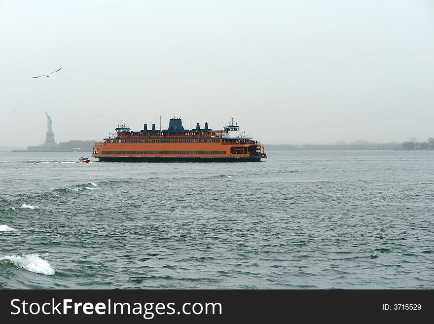 A picture of an Orange Ship in the New York Habor with statue of Liberty in distance background. A picture of an Orange Ship in the New York Habor with statue of Liberty in distance background