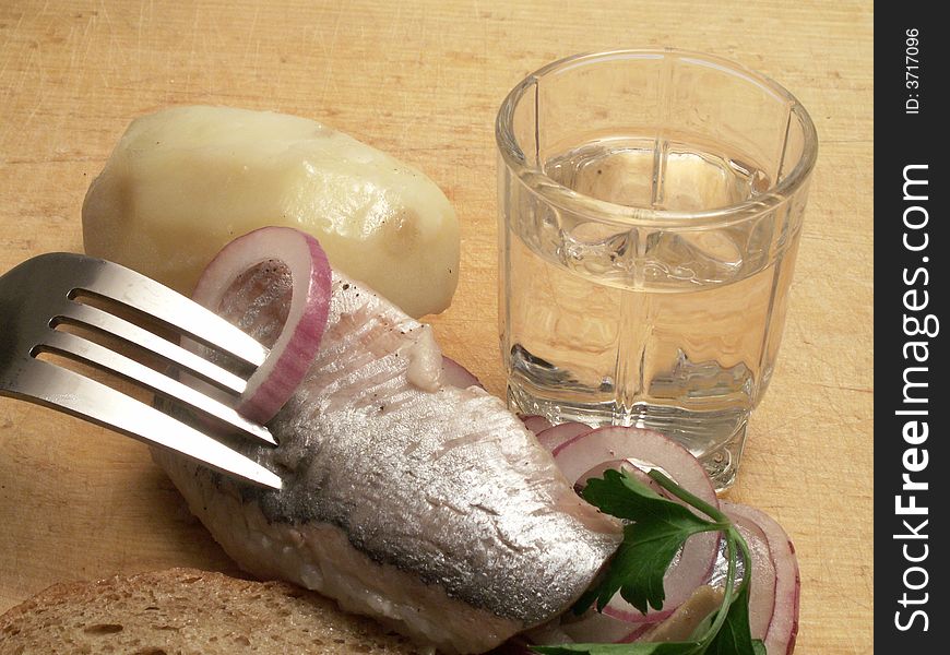 Glass of vodka, boiled potatoes and fork with herring and onion on background with wooden plate. Glass of vodka, boiled potatoes and fork with herring and onion on background with wooden plate