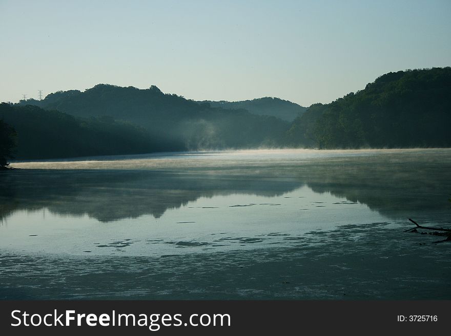 The sun rises as steam comes off a scenic lake with hills reflected in the distance. The sun rises as steam comes off a scenic lake with hills reflected in the distance.