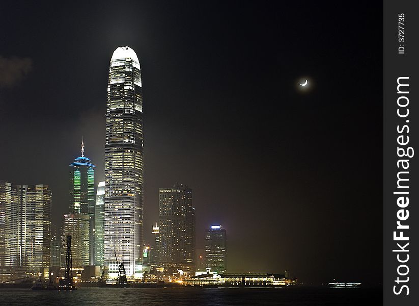 Central Business district of Hong Kong at night with a crescent moon. Ferry piers and victoria harbor ( harbour ) in the foreground, with the office buildings and skyscrapers of the city behind. Central Business district of Hong Kong at night with a crescent moon. Ferry piers and victoria harbor ( harbour ) in the foreground, with the office buildings and skyscrapers of the city behind.