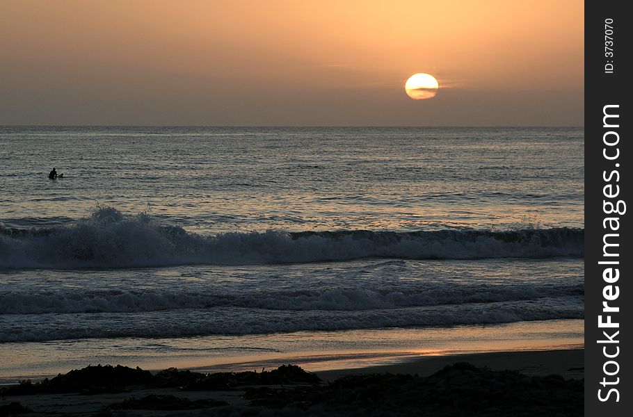 This is a photo taken on a Southern California beach.  There is a surfer sitting on his board watching the sunset from the water. This is a photo taken on a Southern California beach.  There is a surfer sitting on his board watching the sunset from the water.