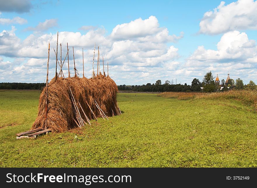 A hayfield with a haystack near the village named Pogost in the north of Russia. There is an old wooden church in the background. A hayfield with a haystack near the village named Pogost in the north of Russia. There is an old wooden church in the background.