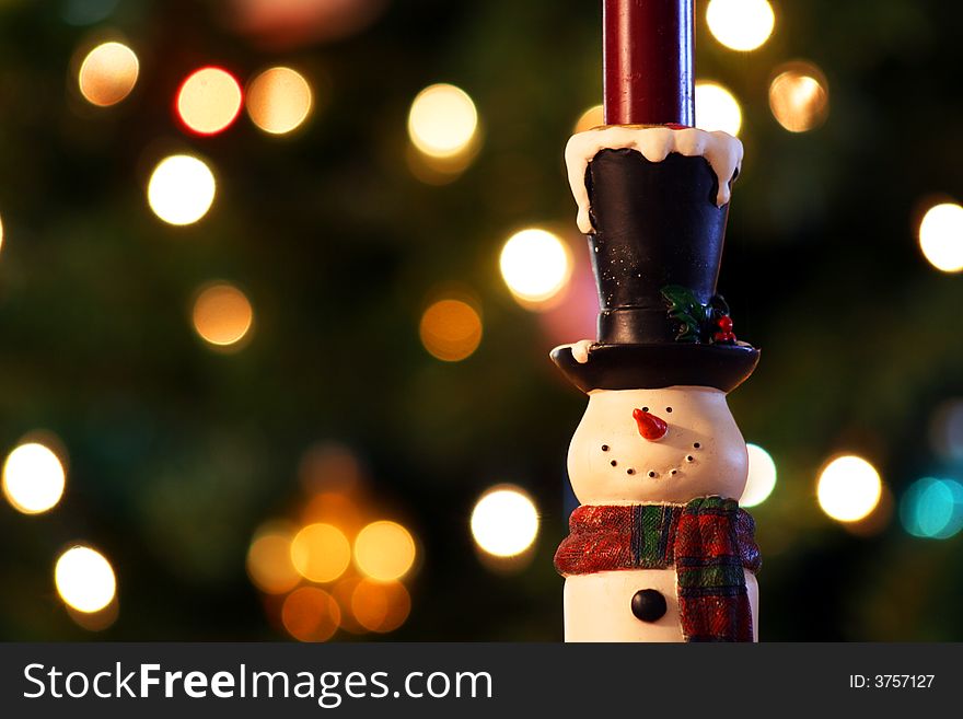 A cheerful snowman candle holder stands before the colored lights of a Christmas tree. A cheerful snowman candle holder stands before the colored lights of a Christmas tree