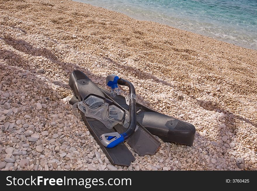 Photo of swimming flippers, mask and respiratory tube lying on pebble near water. Photo of swimming flippers, mask and respiratory tube lying on pebble near water