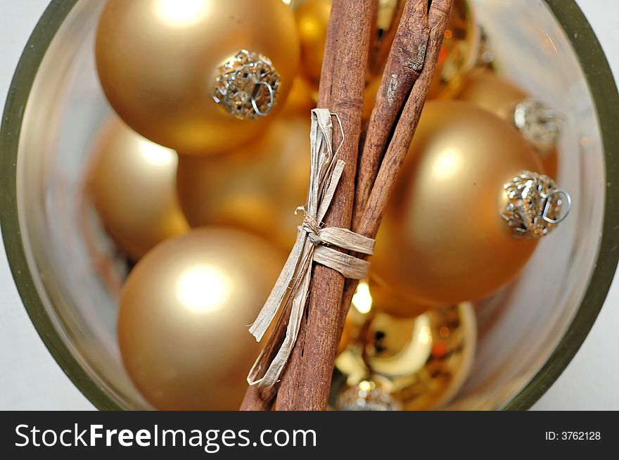 A closeup of golden Christmas balls, in a glass container with a bunch of cinnamon sticks placed across the mouth of the glass. A closeup of golden Christmas balls, in a glass container with a bunch of cinnamon sticks placed across the mouth of the glass.