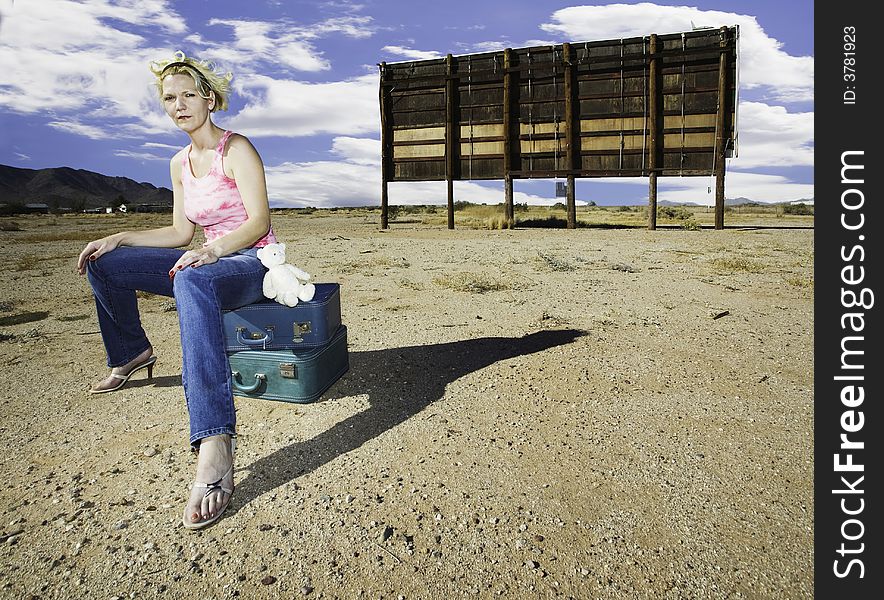 Woman sitting on suitcases in front of an old billboard waiting in the desert. Woman sitting on suitcases in front of an old billboard waiting in the desert.