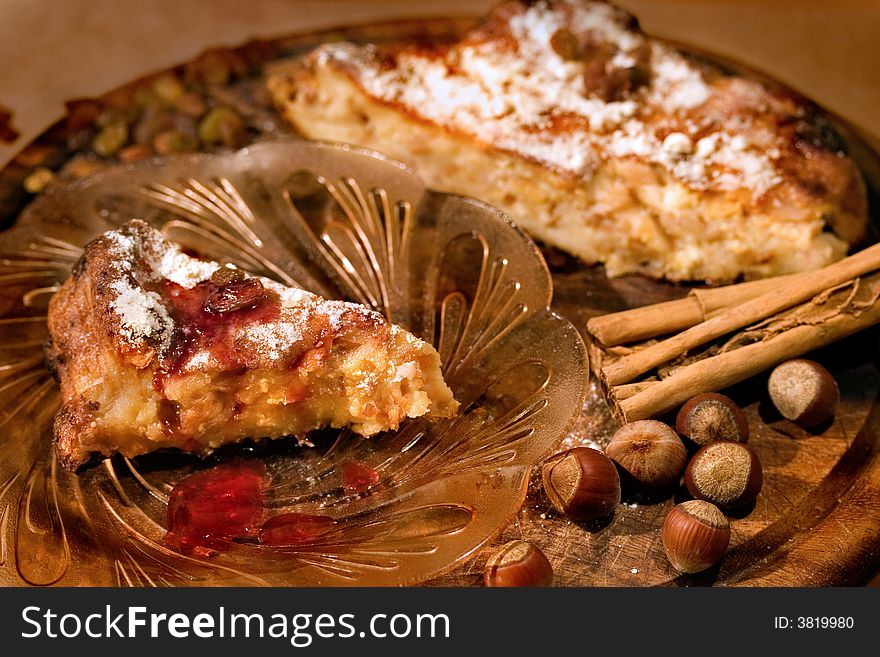 A slice of delicious homemade cake in front on the glass plate. The cake itself is in the backround together with cinnamon sticks, filbert nuts and raisins. Tha focus is on the slice of cake and nuts in the foreground.