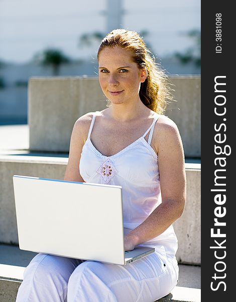 Woman (a student?) sitting alone on a stair, typing on the silver laptop. Looking friendly to you. Bright and summery. Vertical orientation. Woman (a student?) sitting alone on a stair, typing on the silver laptop. Looking friendly to you. Bright and summery. Vertical orientation.