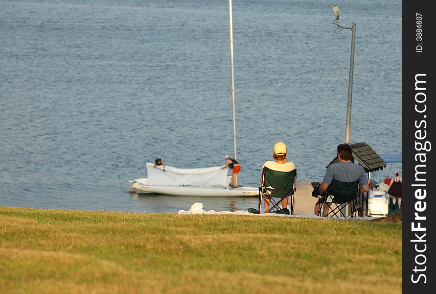 A couple sit in lawn chairs and watch other people prepare a sailboat for a day on the lake. A couple sit in lawn chairs and watch other people prepare a sailboat for a day on the lake.