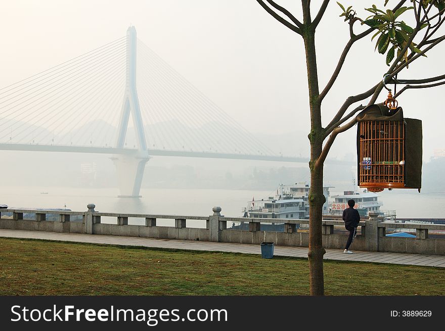 A Birdcage is hanged by Branches,nearby,a women is doing Physical Exercise in the edge of Yangtze River in the morning. A Birdcage is hanged by Branches,nearby,a women is doing Physical Exercise in the edge of Yangtze River in the morning.