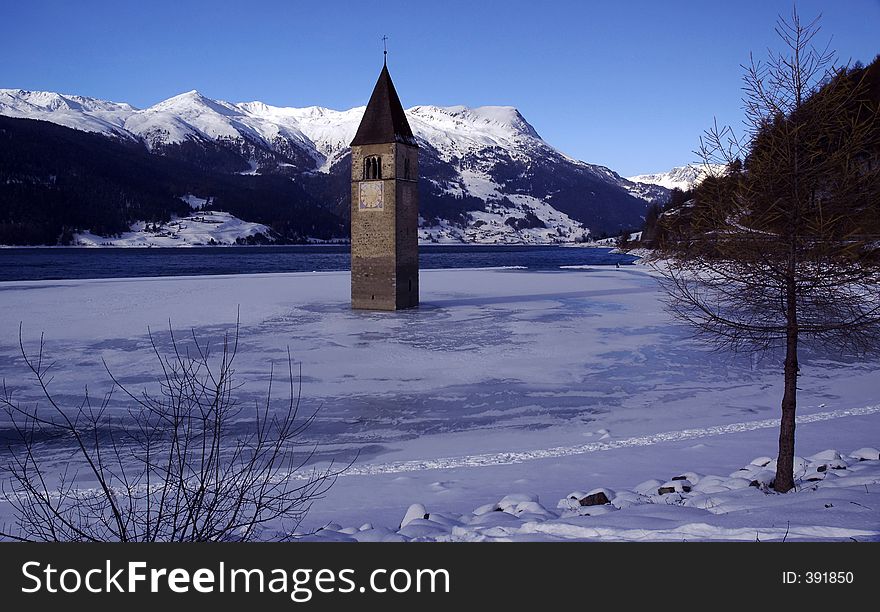 It shows a a bell tower of a church which is standing in a lake with view to Austria