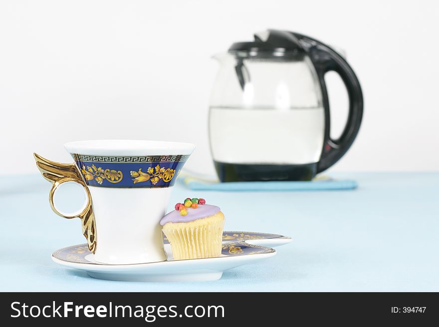 A cup of coffee or tea and a mini cupcake. Kettle in background out of focus. A cup of coffee or tea and a mini cupcake. Kettle in background out of focus.