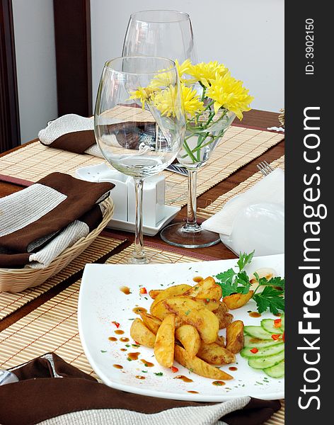 The Fried potatoes and cut vegetables on plate in restaurant. On back background tall wine glasses.