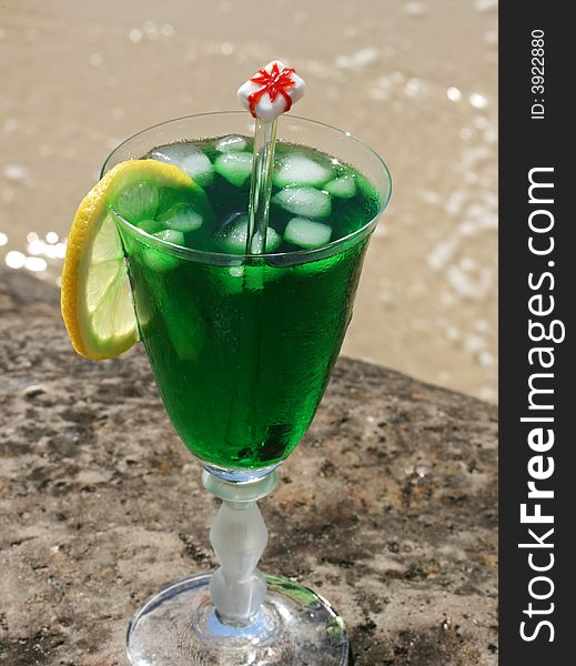 Green Christmas cocktail on a rock at the beach, with the ice dropped in it, with the beach and waves in the background. Green Christmas cocktail on a rock at the beach, with the ice dropped in it, with the beach and waves in the background