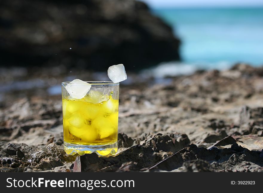 Tumbler glass with ice falling into the glass containing whiskey with the ocean and blue sky in the background. Tumbler glass with ice falling into the glass containing whiskey with the ocean and blue sky in the background