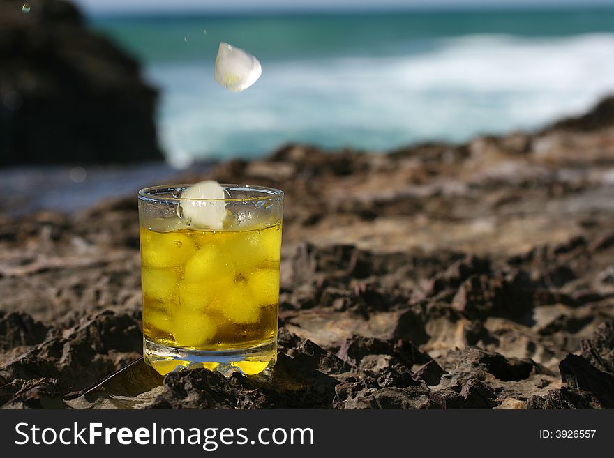 Tumbler glass with ice falling into the glass containing whiskey with the ocean and blue sky in the background. Tumbler glass with ice falling into the glass containing whiskey with the ocean and blue sky in the background