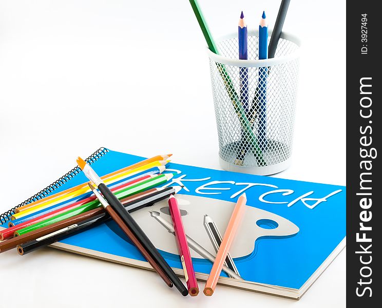 Still life of a sketch pad, color pencils, paintbrushes and a pencil holder on white background. Still life of a sketch pad, color pencils, paintbrushes and a pencil holder on white background