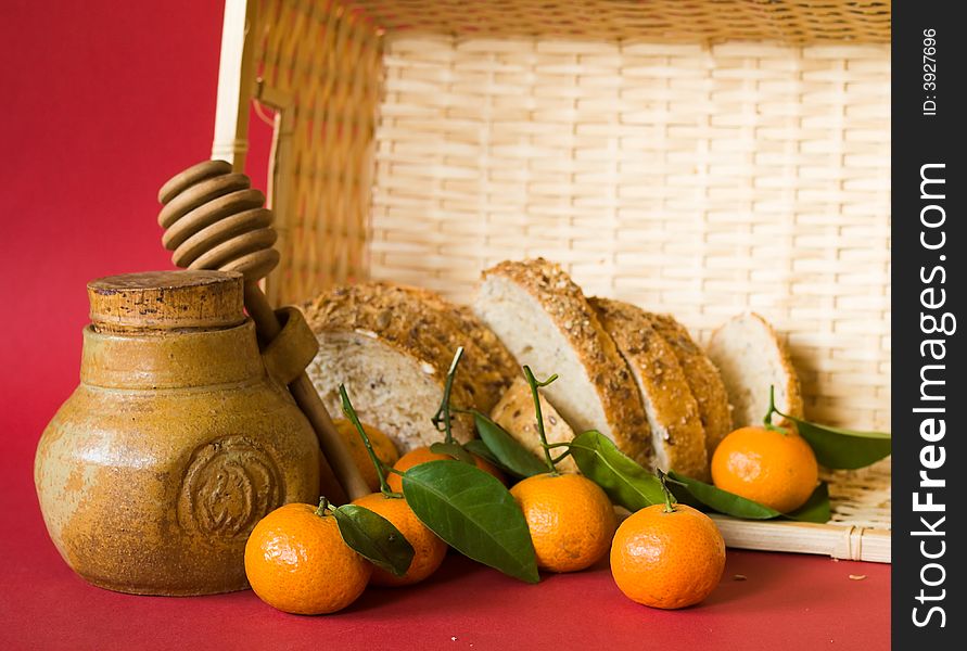Still life of a honey pot with dipper, tangerines  with leaves , and a loaf of bread in a wicker basket, on red background illuminated by the morning sun from the side. Still life of a honey pot with dipper, tangerines  with leaves , and a loaf of bread in a wicker basket, on red background illuminated by the morning sun from the side