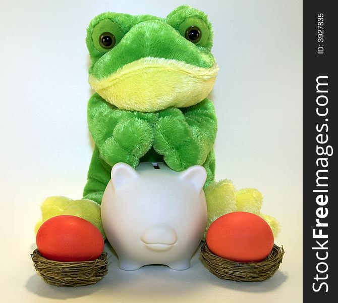 Cute plush frog sitting behind a white ceramic piggy bank with two red Easter eggs in nests. Cute plush frog sitting behind a white ceramic piggy bank with two red Easter eggs in nests.