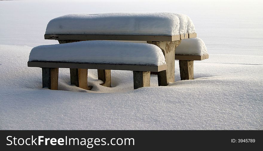 A lakeside picnic table and benches are covered with snow. A lakeside picnic table and benches are covered with snow.