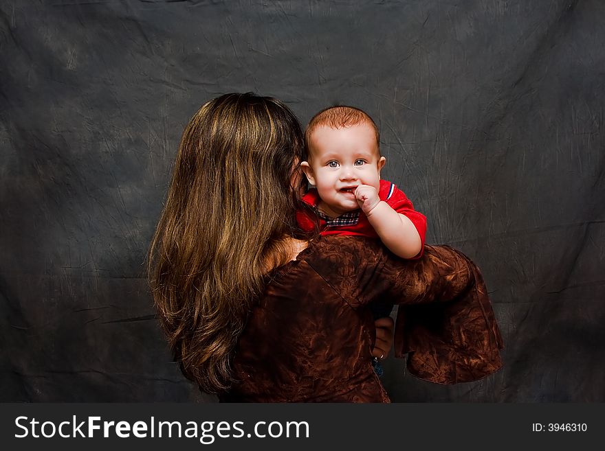 Mother dressed in brown from the back is holding her baby boy with sparkling eyes who's sucking his thumb, having a mischievous look and facing camera, isolated on a dark background. Mother dressed in brown from the back is holding her baby boy with sparkling eyes who's sucking his thumb, having a mischievous look and facing camera, isolated on a dark background