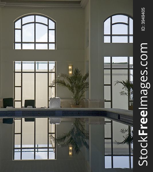 Lounge chairs and potted palm tree reflected with windows in a quiet indoor pool in a spa. Lounge chairs and potted palm tree reflected with windows in a quiet indoor pool in a spa