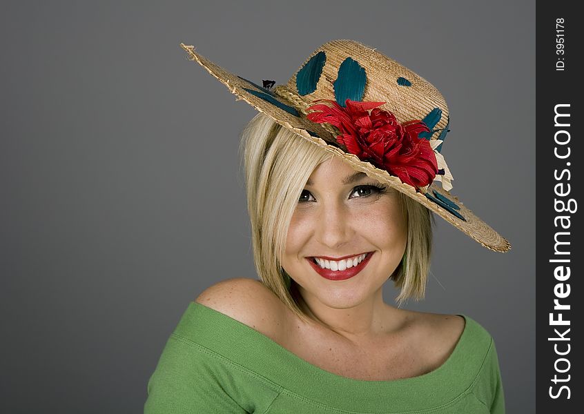 A blonde model with brilliant smile in a flowered straw hat on grey background. A blonde model with brilliant smile in a flowered straw hat on grey background