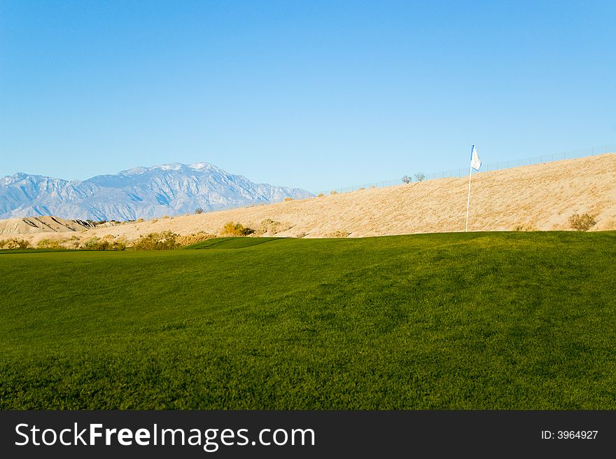 An image of a golf course in a desert with a flag in a foreground and mountains in the background.