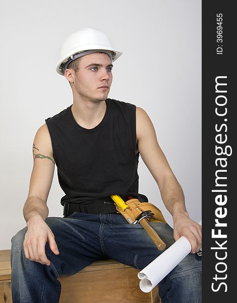 Young man wearing hardhat and toolbelt, sitting on a wooden crate and holding a set of blueprints. Young man wearing hardhat and toolbelt, sitting on a wooden crate and holding a set of blueprints.