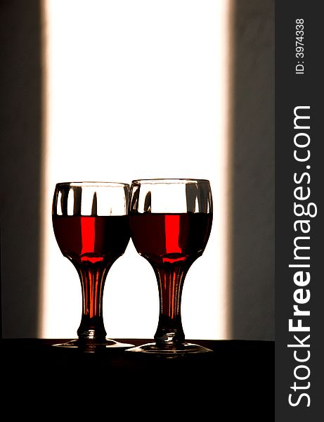 Wine glasses stand with a bottle of wine dramatically backlit to show of the beauty of the color of the containersâ€™ contents. Wine glasses stand with a bottle of wine dramatically backlit to show of the beauty of the color of the containersâ€™ contents.