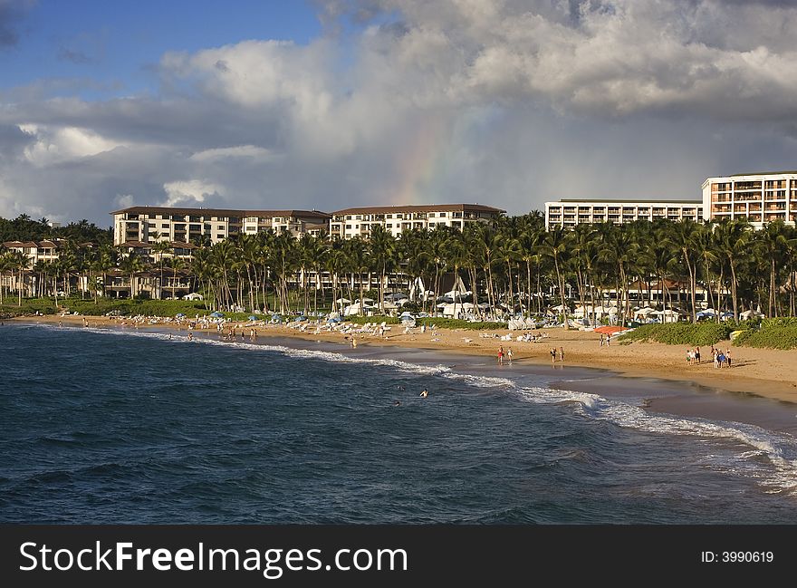 A small rainbow crowns hotels along a palm tree lined golden beach and blue ocean. A small rainbow crowns hotels along a palm tree lined golden beach and blue ocean.