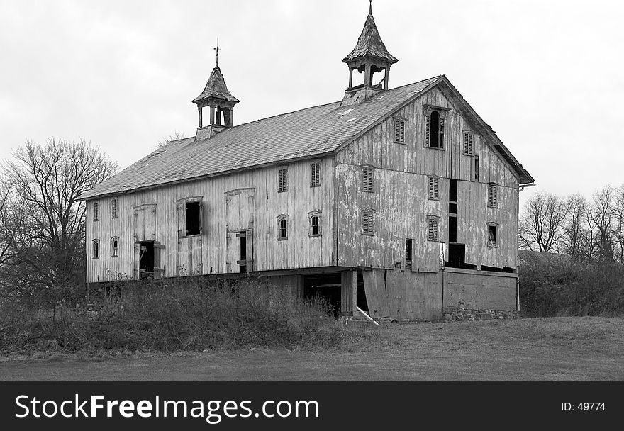 A large, very old barn in disrepair photographed in black and white. Many boards are missing and shingles fall from the roof as the barn slowly over the years decays. Of interest are the steeples on the roof, symbols of a successful farm in the days when this barn was first built. A large, very old barn in disrepair photographed in black and white. Many boards are missing and shingles fall from the roof as the barn slowly over the years decays. Of interest are the steeples on the roof, symbols of a successful farm in the days when this barn was first built