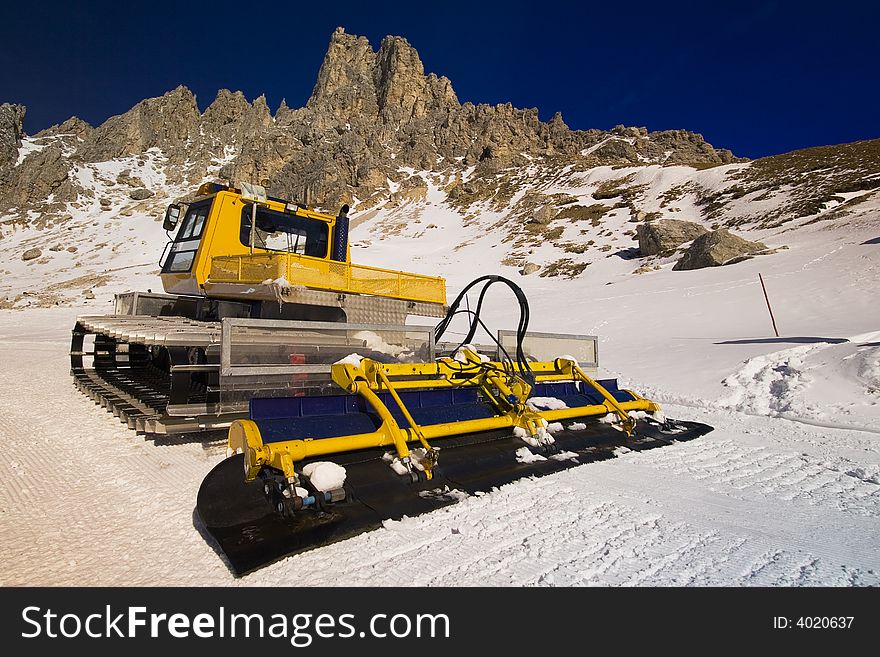 A snow plough on a mountain ski slope with jagged peaks in the background. A snow plough on a mountain ski slope with jagged peaks in the background.