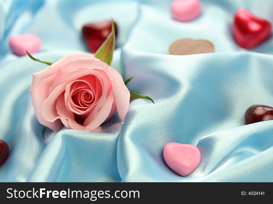 A pink rose surrounded by little hearts of chocolate, jelly and other materials lying on blue satin fabric. A pink rose surrounded by little hearts of chocolate, jelly and other materials lying on blue satin fabric.