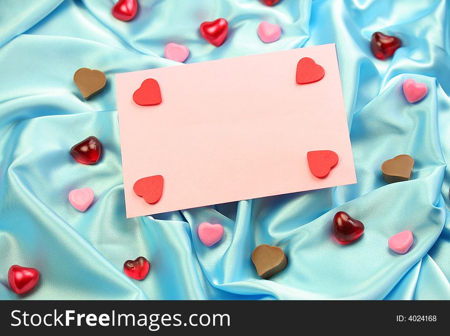 A pink valentine card lying on blue satin fabric with little hearts of different materials.
