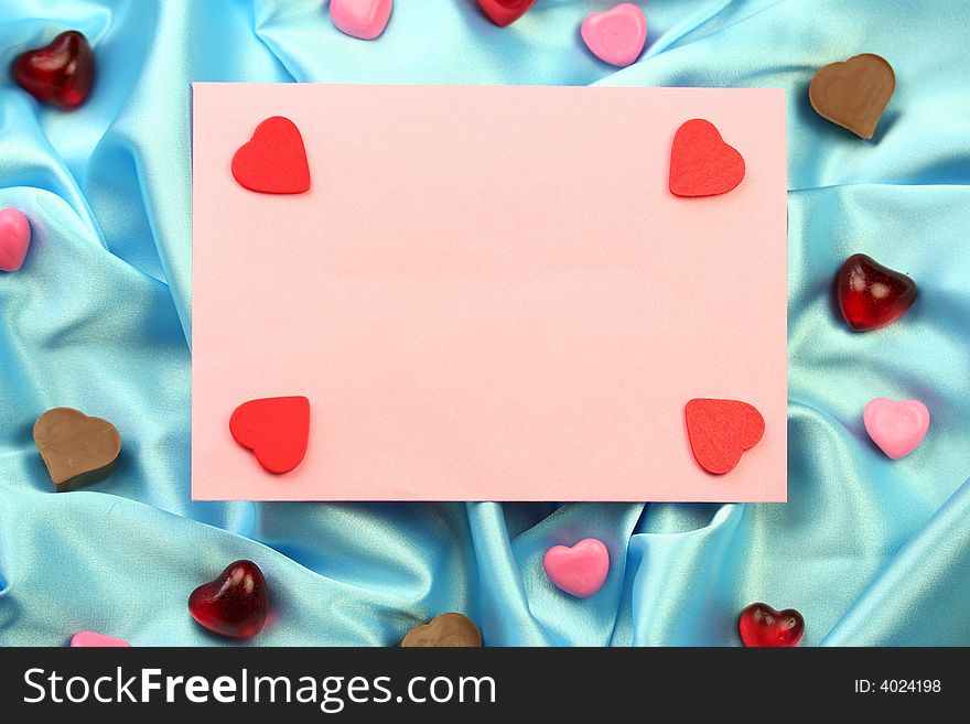 A pink valentine card lying on blue satin fabric with little hearts of different materials. A pink valentine card lying on blue satin fabric with little hearts of different materials.