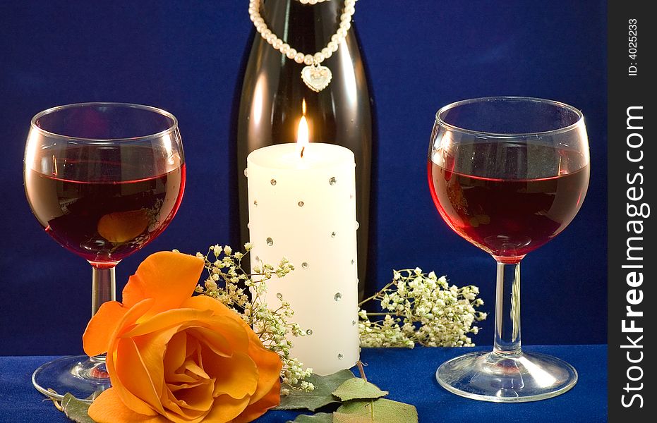 A closeup view of two glasses filled with red wine, a white candle and a peach rose with a bottle of wine and baby's breath in the background. A closeup view of two glasses filled with red wine, a white candle and a peach rose with a bottle of wine and baby's breath in the background.