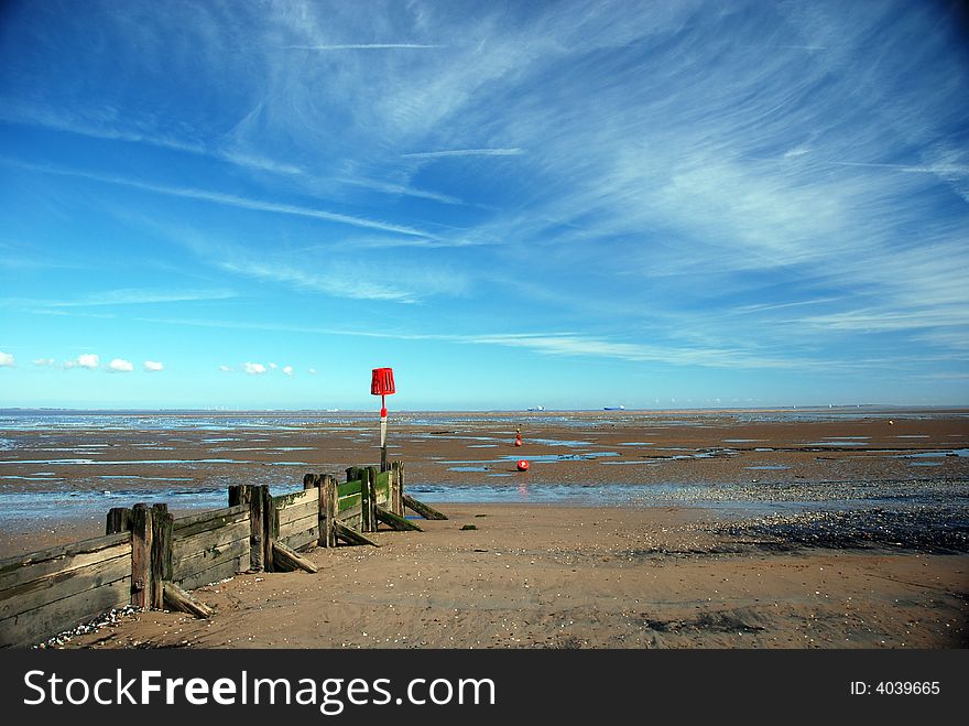 Estuary beach at low tide showing very large area of sand, groyne or breakwater, and red danger marker set against dramatic sky showing high clouds and con trails