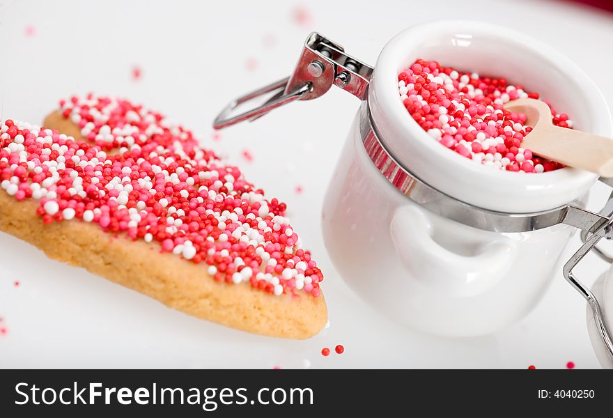 Red, white and pink nonpareils on heart-shaped cookie and in white porcelin storage jar. Red, white and pink nonpareils on heart-shaped cookie and in white porcelin storage jar.