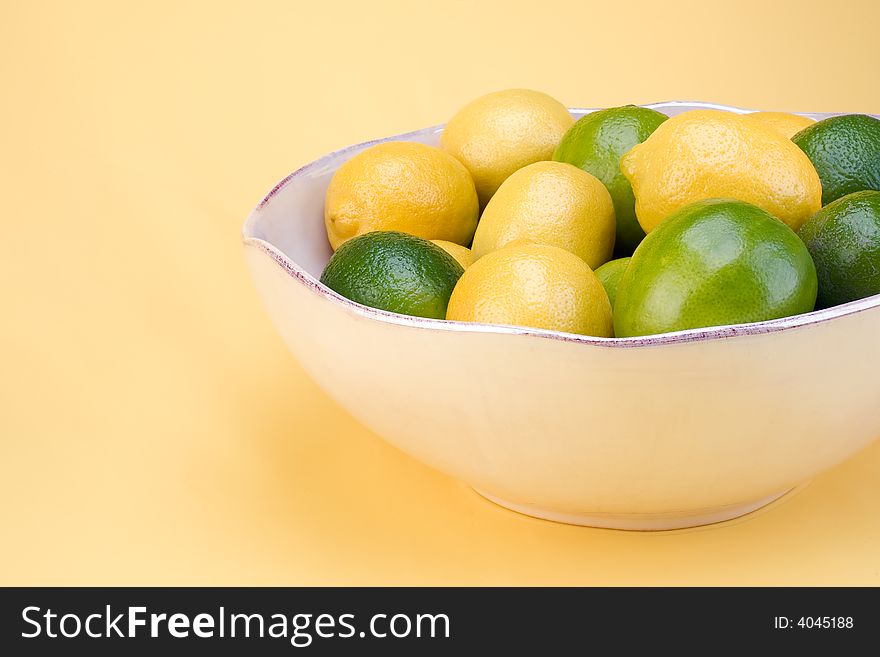 A bowl of lemons and limes in a white bowl on a yellow background. A bowl of lemons and limes in a white bowl on a yellow background.