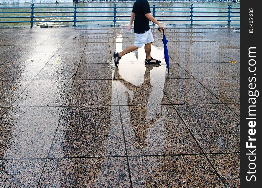 A young man strolling along the wet walkway by a river with an umbrella after a late afternoon shower. A young man strolling along the wet walkway by a river with an umbrella after a late afternoon shower