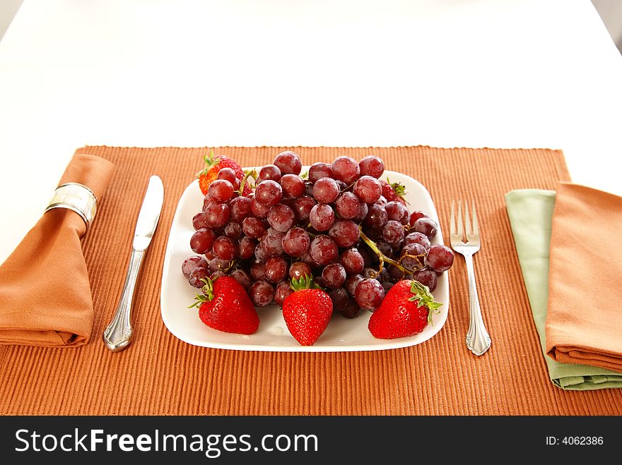 Concept: Eat more fruit. Plate piled high with red grapes, accented with strawberries, napkins, fork, and table accessories. Concept: Eat more fruit. Plate piled high with red grapes, accented with strawberries, napkins, fork, and table accessories.