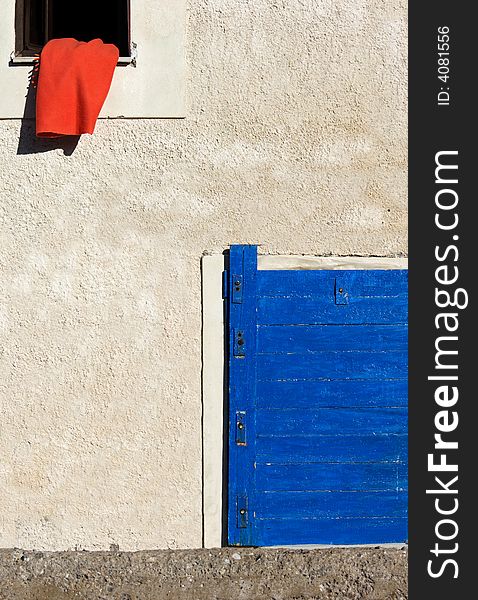 Image shows a red blanket on the window of a Greek village house. A blue door appears in the lower right corner. Image shows a red blanket on the window of a Greek village house. A blue door appears in the lower right corner