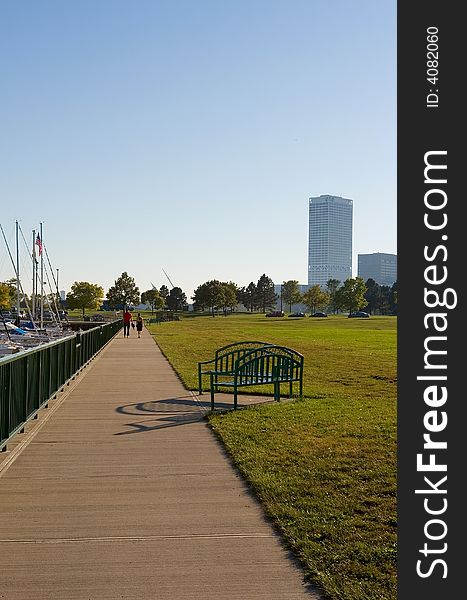 Sidewalk along marina and park with two people strolling in the distance and buildings on the horizon. Sidewalk along marina and park with two people strolling in the distance and buildings on the horizon