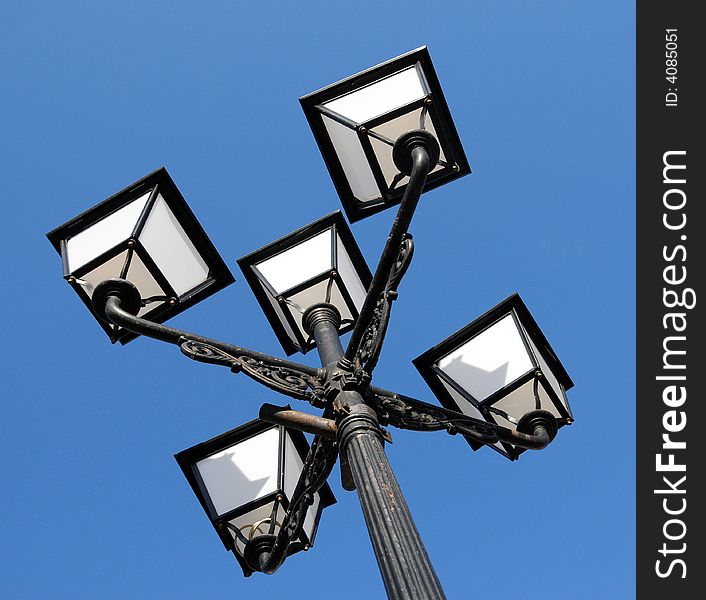 Three ornate street lamps on a lamp post against a blue sky in Romania