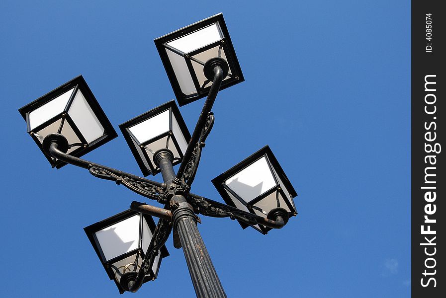 Three ornate street lamps on a lamp post against a blue sky in Romania