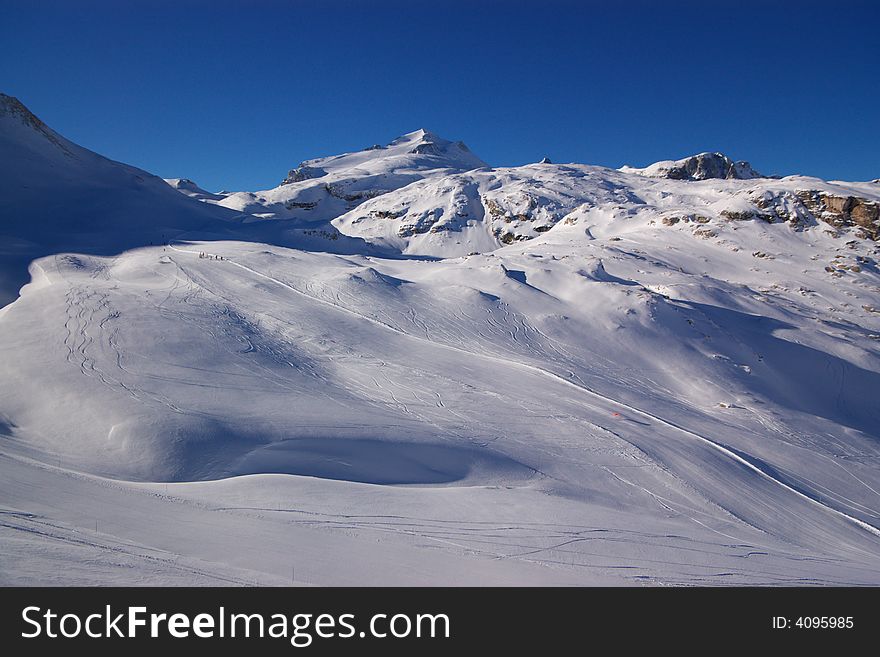 View on alpine snow winter mountains chain and downhill slopes under blue sky. Alps, France, Europe. View on alpine snow winter mountains chain and downhill slopes under blue sky. Alps, France, Europe.