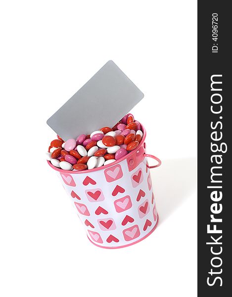 A bucket full of red, white and pink candies with a blank grey card to add message with copy space on a white background. A bucket full of red, white and pink candies with a blank grey card to add message with copy space on a white background